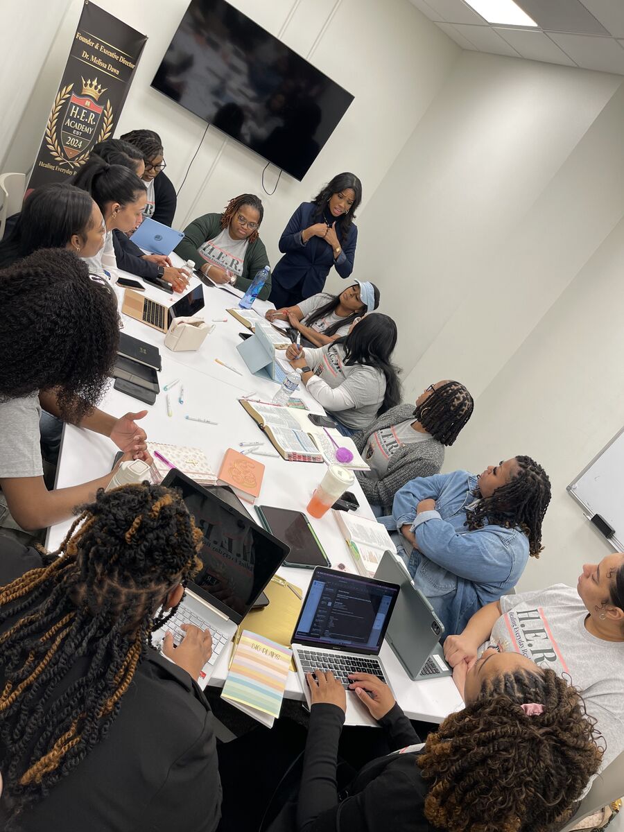 Women seated around a long conference table with laptops and workbooks during a group coaching session