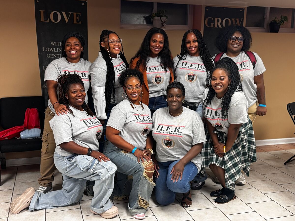 A group of ten women in H.E.R. Academy shirts smiling together beneath wall art reading LOVE and GROW