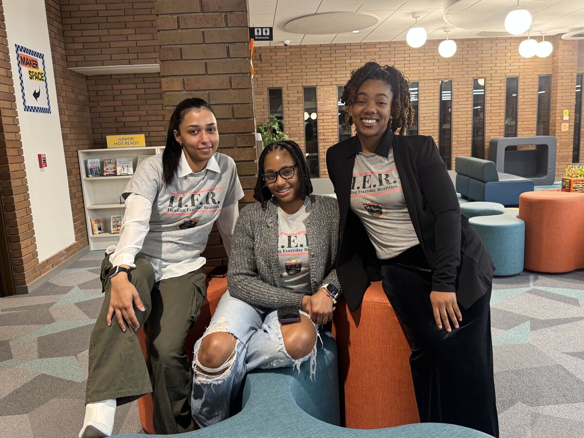 Three H.E.R. Academy members in branded shirts sitting together and laughing in a bright lounge
